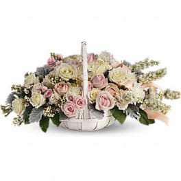 Low white basket filled with pastel pink and cream roses and small white blossoms.