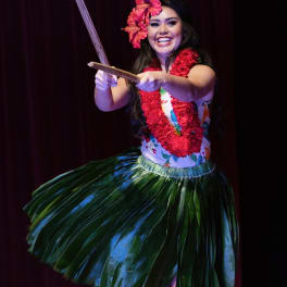 Hula dancer wearing a red flower lei and hair flower with a green grass skirt on stage