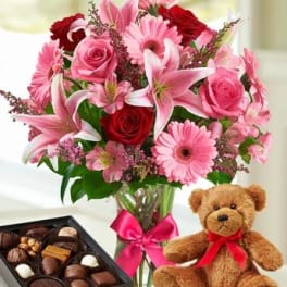 Pink and red flower bouquet in a glass vase with a teddy bear and chocolates