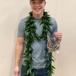 Person wearing a long green leaf lei and holding a small wrapped congratulations gift.