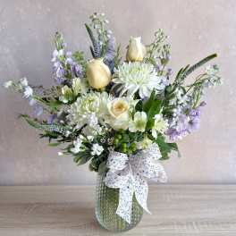 Pastel arrangement of white mums, cream roses, and lavender blooms in a textured glass vase with a ribbon bow.