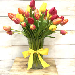 Mixed red, yellow, and orange tulips in a clear glass vase with a yellow ribbon bow.