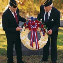 Patriotic standing wreath of white flowers with red, white, and blue accents and ribbon on an easel outdoors.