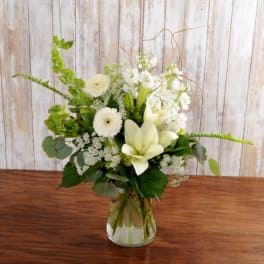 Tall white and green flower arrangement with lilies and gerbera daisies in a clear glass vase on a wooden table.