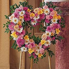 Heart-shaped standing wreath of pink, yellow, and white flowers on a gold easel.