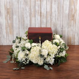 Wooden cremation urn surrounded by white hydrangeas, carnations, and mums on a table.