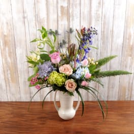 Tall mixed bouquet of pink roses, tulips, hydrangeas, and purple blooms in a white ceramic pitcher vase