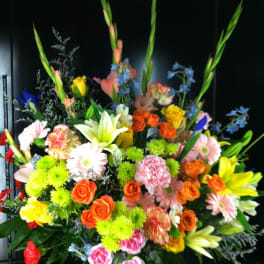 Tall, colorful mixed flower arrangement with roses, lilies, gerbera daisies and carnations in a patterned vase.