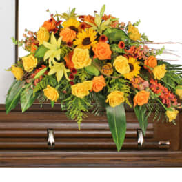 Yellow and orange casket spray of roses, lilies, and sunflowers on a wooden casket.