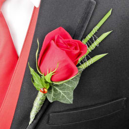 Single red rose boutonniere with decorative green blades on a black suit with red vest.