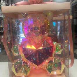 A faceted iridescent teddy bear in a clear gift box with a pink ribbon