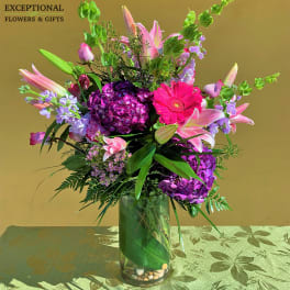 Tall arrangement of pink lilies, purple hydrangeas, and bright pink gerbera in a clear glass vase.