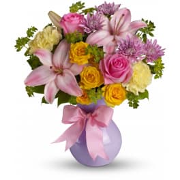 Bouquet of pink lilies, roses, and purple flowers in a lavender vase with a pink ribbon