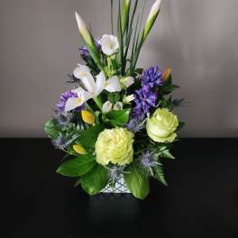 Mixed bouquet with white lilies, purple blooms, and pale green roses in a square vase