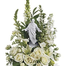 White roses and lilies arranged around a white religious figurine with tall white blooms.