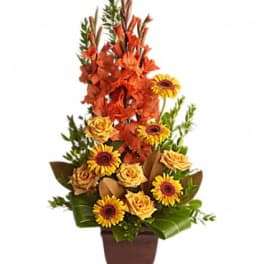 Tall orange and yellow flower arrangement with roses and daisy-like blooms in a brown pedestal vase