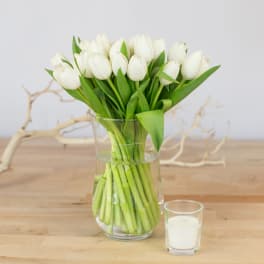 White tulips in a clear glass vase beside a small white candle