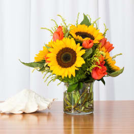 Yellow sunflowers and orange roses in a clear glass vase on a table with a white seashell.