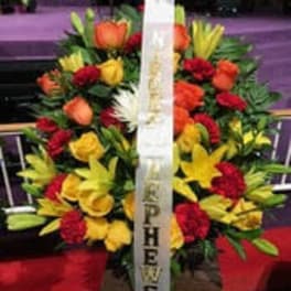 Standing funeral spray of red, yellow, and orange flowers with a white ribbon