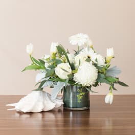 Low arrangement of white flowers in a glass vase beside a large white seashell on a wooden surface.
