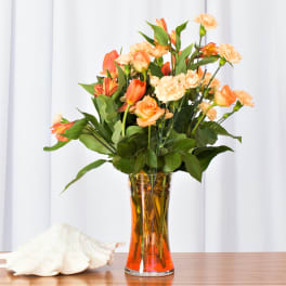 Tall arrangement of orange and peach roses, carnations, and other blooms in a clear glass vase with orange water.