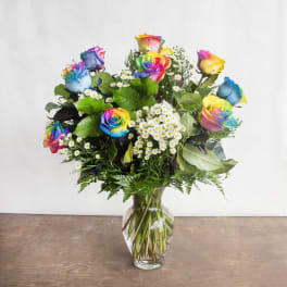 Arrangement of rainbow-colored roses and small white daisies in a clear glass vase on a wooden surface.