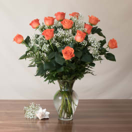 Tall arrangement of orange roses and white filler flowers in a clear glass vase on a wooden table.