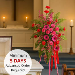 Bright pink and red standing spray on an easel beside a white casket in a chapel.