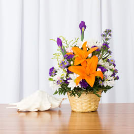 Basket arrangement of orange lilies, purple flowers, and white daisies beside a large seashell.