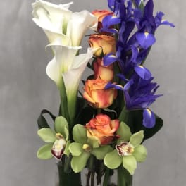 Tall arrangement of white calla lilies, orange roses, and purple irises in a glass vase