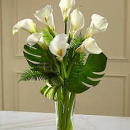 White calla lilies arranged in a clear glass vase with large tropical leaves