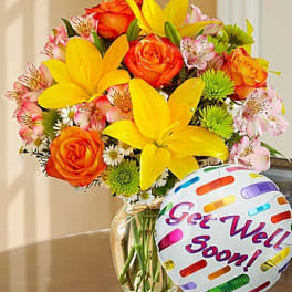 Bouquet of yellow lilies and orange roses in a glass vase with a "Get Well Soon" balloon