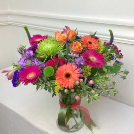 Vibrant mixed bouquet of orange roses and gerbera daisies with purple and green blooms in a clear glass vase
