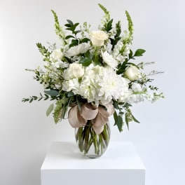 Tall white floral arrangement with roses and mums in a clear glass vase with a beige bow.