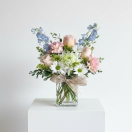 Pastel bouquet of roses, daisies, carnations, and delphinium in a clear glass cube vase with a ribbon bow.