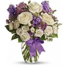 White roses and purple flowers arranged in a clear glass vase with a purple ribbon.