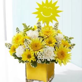 Yellow and white flower arrangement in a square glass vase with a smiling sun topper