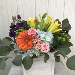 Mixed bouquet in a white wooden box with pink roses, orange gerbera, yellow lilies, and purple blooms