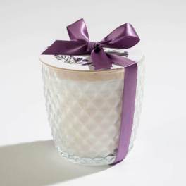 White candle in a textured glass jar with a floral lid and purple satin ribbon bow.