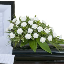 White carnation casket spray with broad green foliage on a black casket beside a white interior lining.