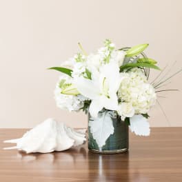 White lilies, roses, and hydrangeas in a glass vase beside a large white seashell.