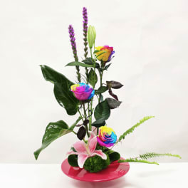 Modern arrangement with rainbow roses, pink lily, and purple spikes in a low pink dish