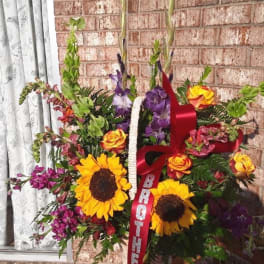 Standing floral spray with sunflowers, roses, and purple blooms on a ribboned easel