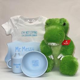 Baby gift set with green dinosaur plush, white onesie, and light blue bib, bowl, and sippy cup.