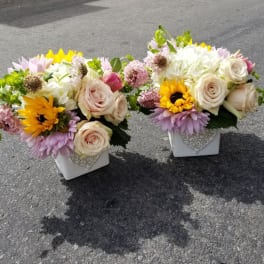 Two floral arrangements in white square vases with roses and sunflowers.