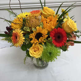 Bright mixed bouquet of yellow roses, sunflowers, red gerberas and green mums in a clear glass vase