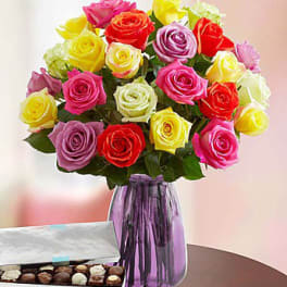 Colorful rose bouquet in a purple vase beside an open box of chocolates.