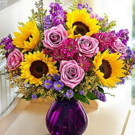 Bouquet of yellow sunflowers, pink roses, and purple flowers in a purple vase