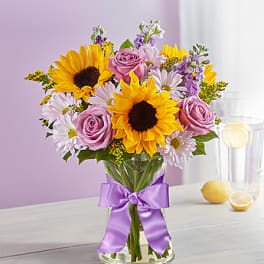 Bouquet of sunflowers, pink roses, and daisies in a glass vase with a purple ribbon