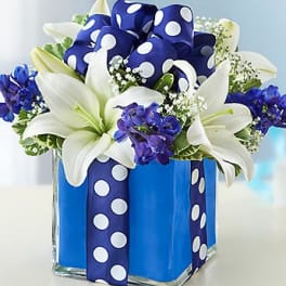 White lilies and blue flowers in a square glass vase with a polka-dot ribbon